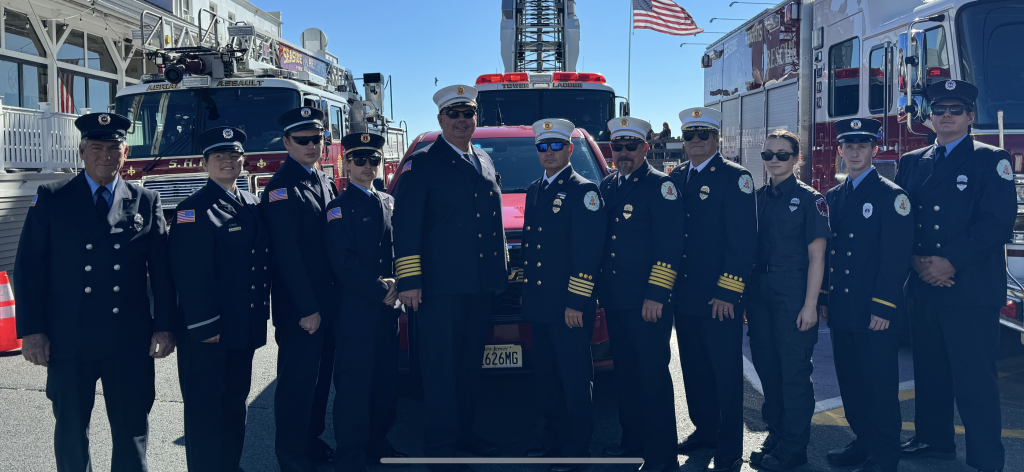 A group of firefighters in uniform stand in a line in front of a fire truck. An American flag is visible in the background. The group includes men and women in various styles of formal fire department attire.