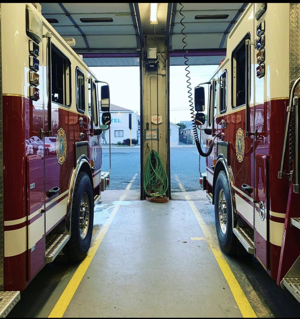 Two red and white fire trucks are parked inside a fire station garage, facing outward. A green hose is coiled on the back wall between the trucks, and the open door reveals a view of the street outside.