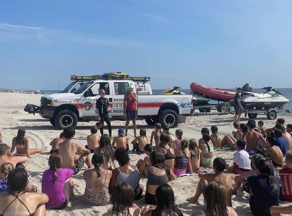 A group of children in swimsuits sit on a sandy beach listening to two lifeguards standing by a rescue truck. Nearby, a jet ski and inflatable rescue boat are visible. The ocean is in the background under a clear blue sky.